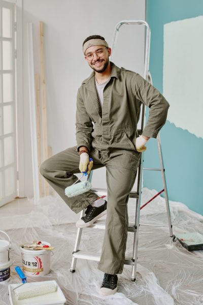 Portrait of young adult man smiling while sitting on ladder holding paint roller, wearing work gloves and coveralls, preparing wall for painting during home renovation